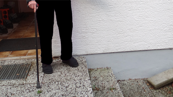 Eine Person mit Gehstock steht neben der Haustür vor zwei nach unten führenden Treppenstufen. (Foto Barrierefrei Leben e.V.)