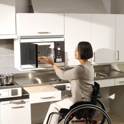 Die Frau im Rollstuhl nutzt Mikrowelle, Oberschränke und Arbeitsplatte mit Liftsystemen zur Höhenverstellung.