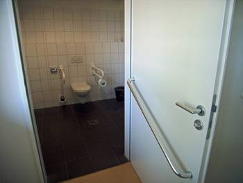 Nach außen aufschlagende Tür in ein Badezimmer mit einer Bedienstange für Rollstuhlfahrer auf der Tür Innenseite.