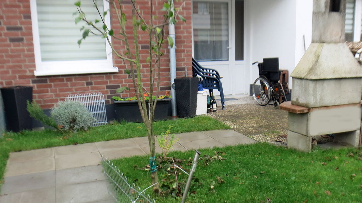 Blick vom Garten auf die Terrasse. Neben der Terassentür, welche als Haupteingang genutzt wird, steht ein Rollstuhl.