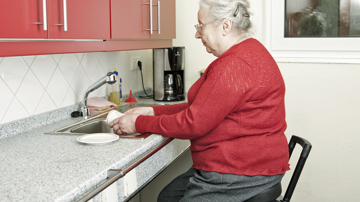Eine Seniorin sitzt vor der Küchenspüle und wäscht ab.