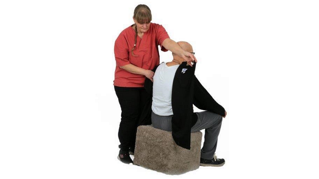 Eine Pflegehelferin zieht einer sitzenden Person ein Oberteil an, das auf der Rückenseite mit Klett geschlossen wird