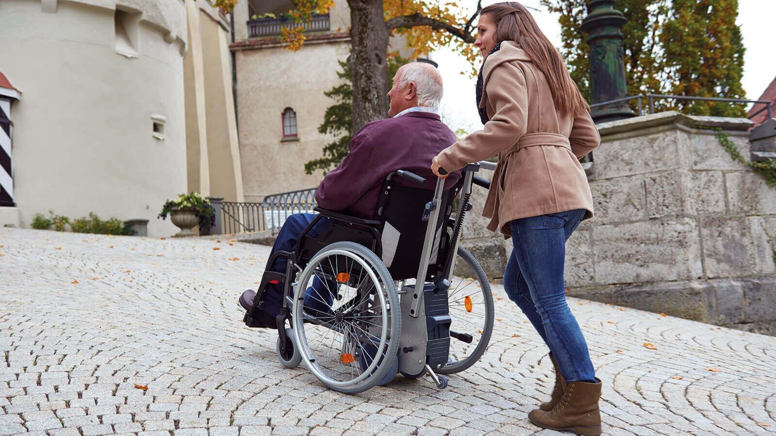 Brems- und Schiebehilfe für manuelle Rollstühle. Ein im Rollstuhl sitzender Mann wird von junger Frau bergauf geschoben.