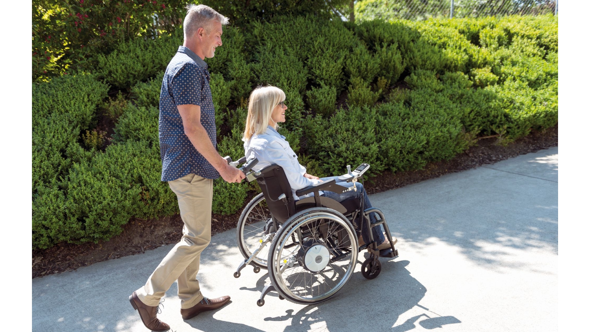 Ein Mann schiebt eine Rollstuhlfahrerin, der Zusatzantrieb hat zwei Steuerungselemente dir von beiden bedient werden können 