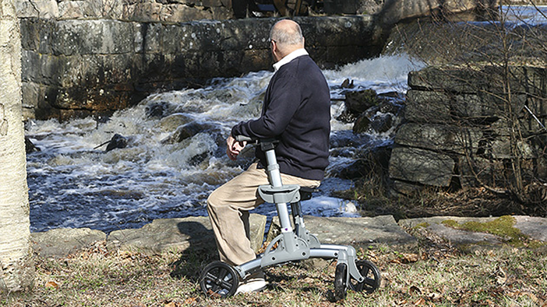Ein Mann sitzt auf einem Gelanderollator vor einem Bergbach.