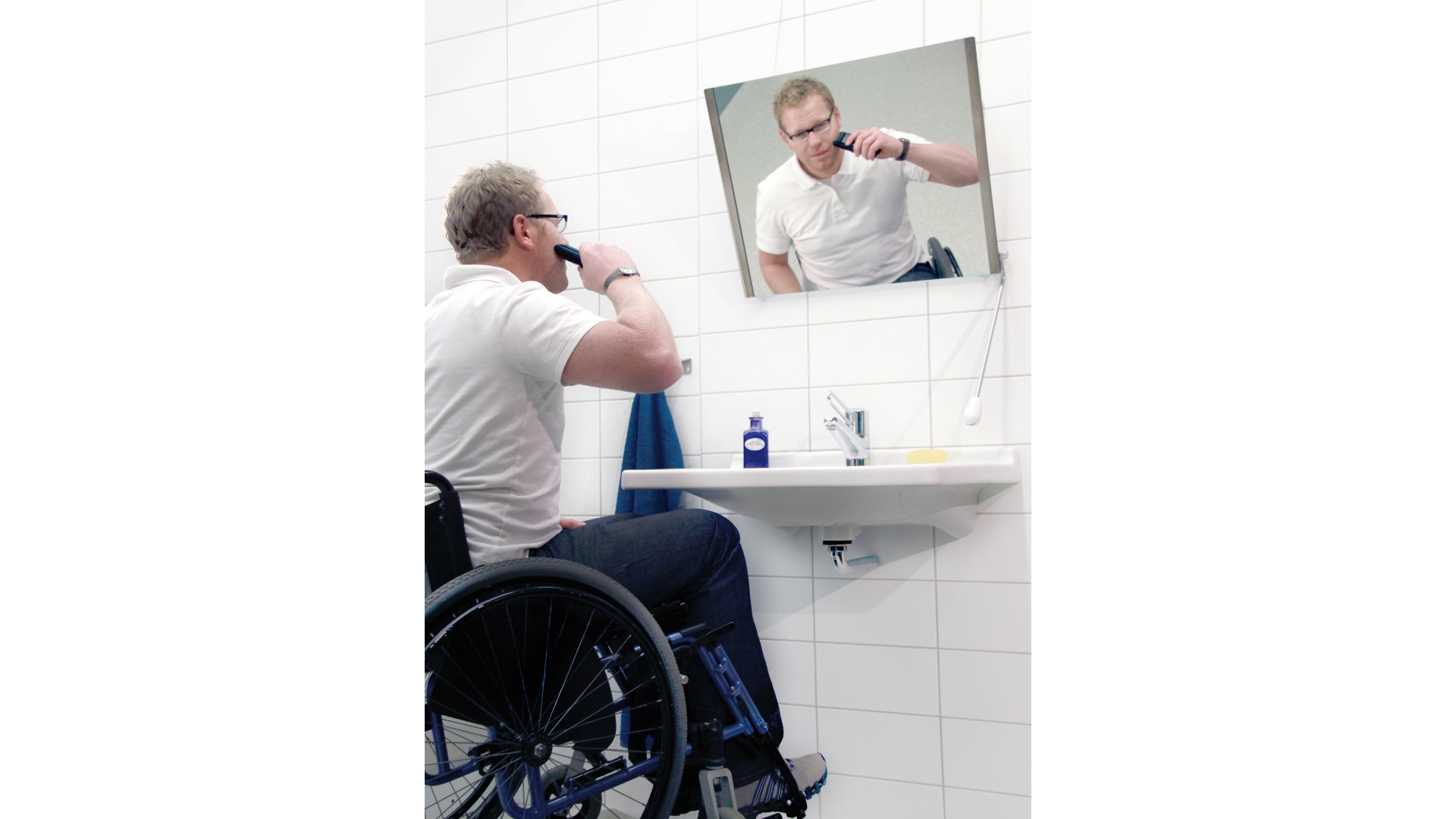 Ein Mann im Rollstuhl steht vor einem Waschbecken und rasiert sich, darüber hängt ein Kippspiegel