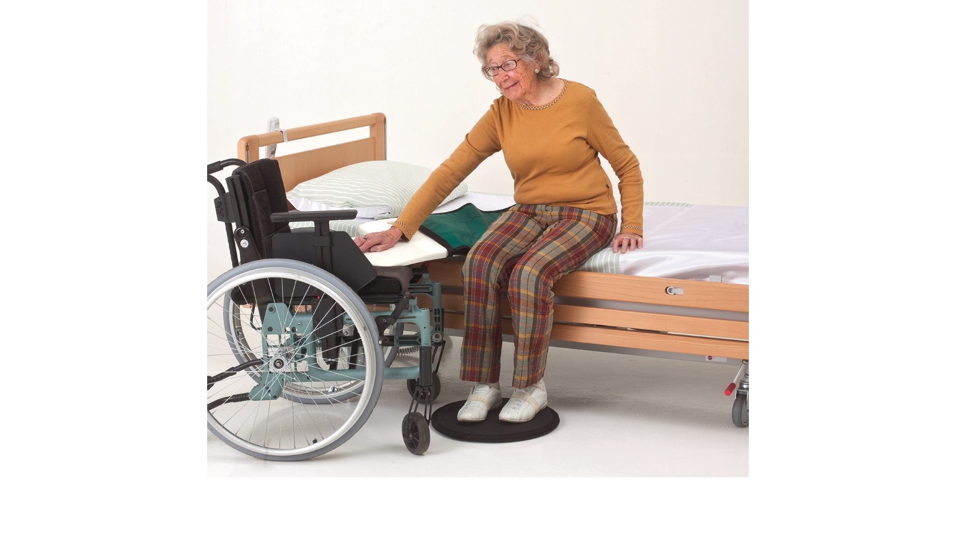 Eine auf der Bettkante sitzende Frau hat ein Rutschbrett von Bettkante zu Rollstuhl gelegt und will sich umsetzen. Unter den Füßen hat sie zur Unterstützung eine Drehscheibe liegen.