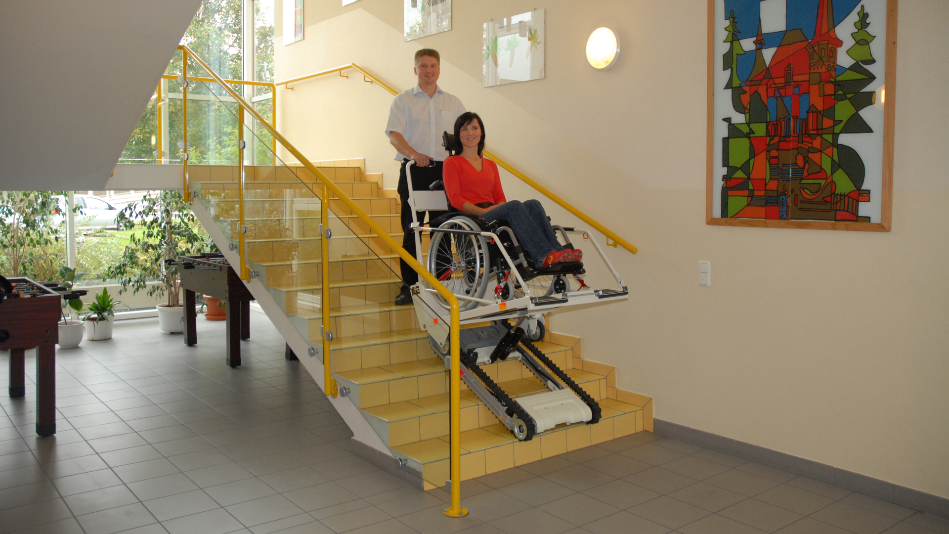 Eine Frau wird sitzend im Rollstuhl mit einer Treppenraupe von einem Mann die Treppe herunter transportiert.