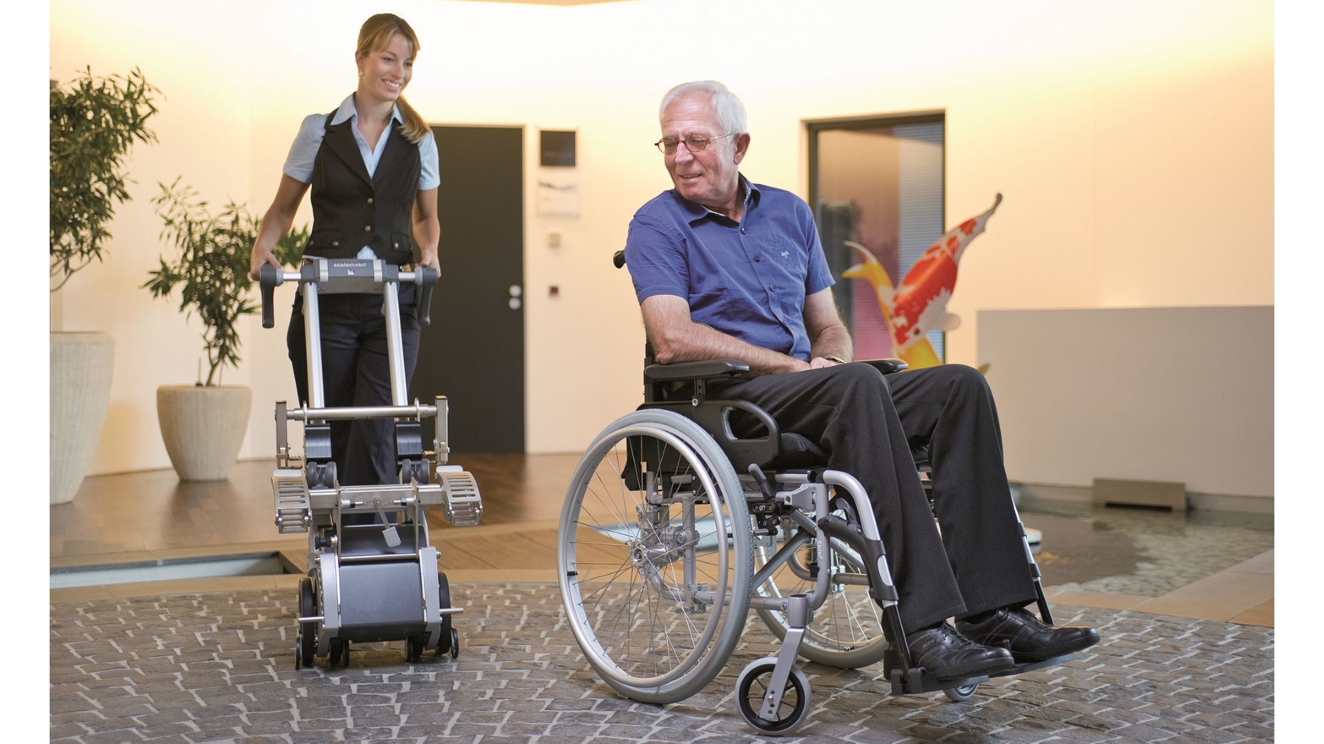 Eine Hilfsperson fährt mit dem Treppensteiger zu einem im Rollstuhl sitzenden Mann.