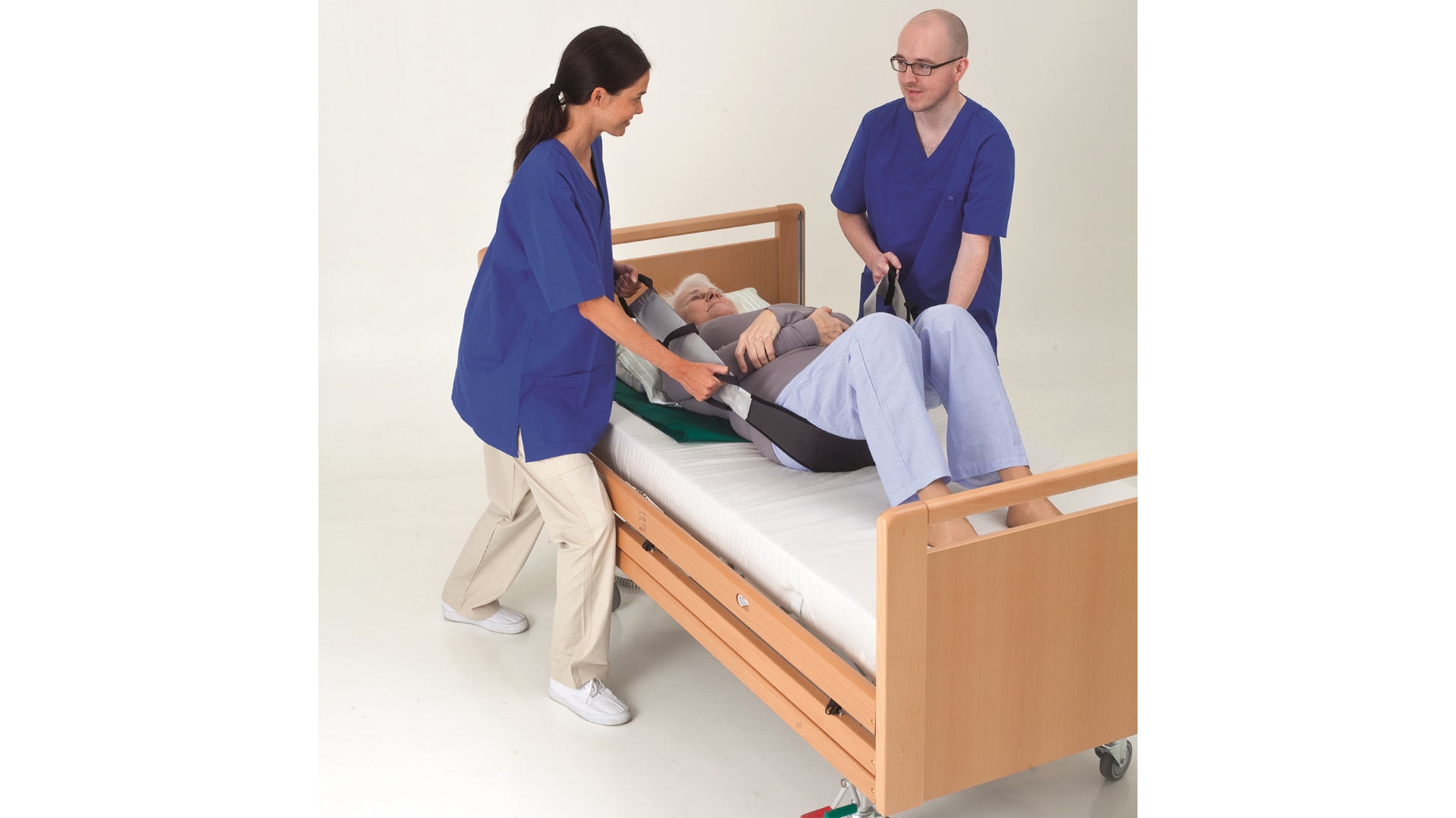 2 Pflegekräfte, männlich und weiblich, stehen auf je einer Bettseite und lagern einen Patienten mit einem Gurt um, der seitliche Griffe hat. 