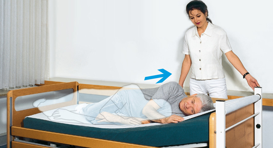 Ein Mann liegt in einem Pflegebett das mit einem beweglichen Bettlaken ausgestattet ist. Per Knopf bewegt die Pflegekraft den Mann in Richtung Kopfteil.