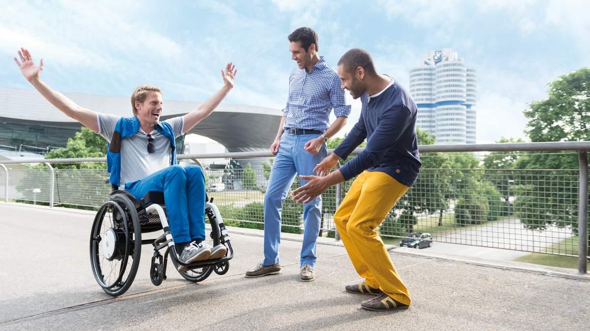 Drei Männer auf einen breiten Fußweg haben Spaß. Der Mann im Rollstuhl balanciert auf zwei Rädern.