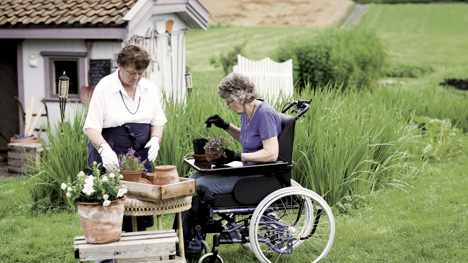 Symbolfoto: Ein Rollstuhlfahrerin und eine stehende Person machen es sich im Gründen an einem gedeckten Tisch gemütlich.
