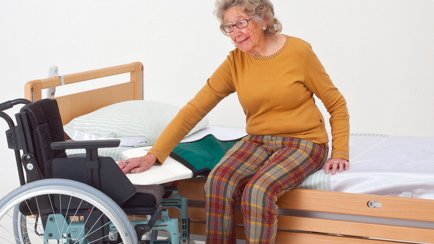 Ein Frau setzt mit Hilfe eines Rutschbrettes vom Bett auf den Rollstuhl um.