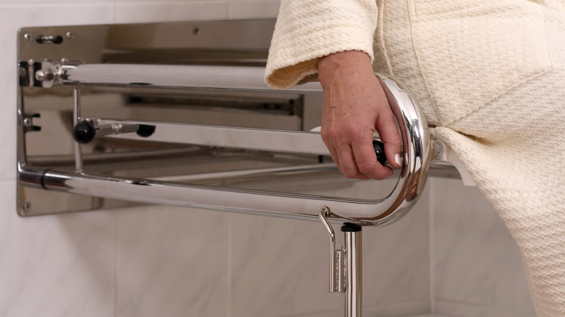 Der Bildausschnitt zeigt die Seitenansicht eines Duschrollstuhls und eine Person im Bademantel.