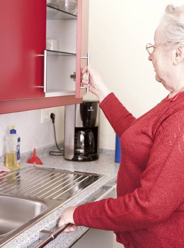 Eine Seniorin öffnet einen Küchenoberschrank, während sie sich an einem Haltegriff festhält, der an der Arbeitsplatte montiert ist. (Ausstellung Barrierefrei Leben e.V.)