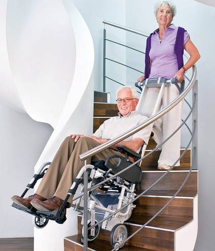 Eine Person im Rollstuhl überwindet mit einem Treppensteiger und einer Hilfsperson eine Treppe.