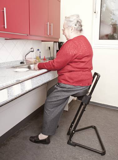 Seniorin auf einer Stehhilfe sitzend, während sie den Abwasch erledigt.(Ausstellung Barrierefrei Leben e.V.)