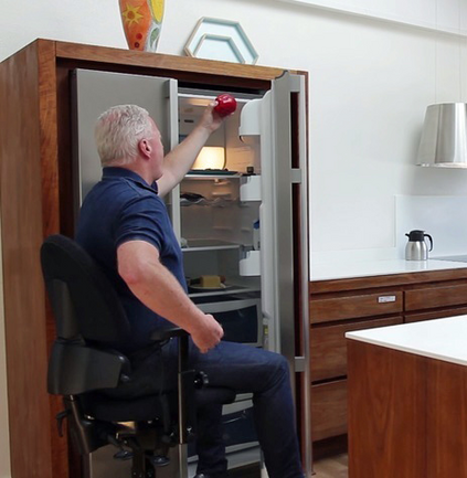 Ein Mann greift sitzend auf einen höhenverstellten Arbeitsstuhl Geschirr in einen Schrank.