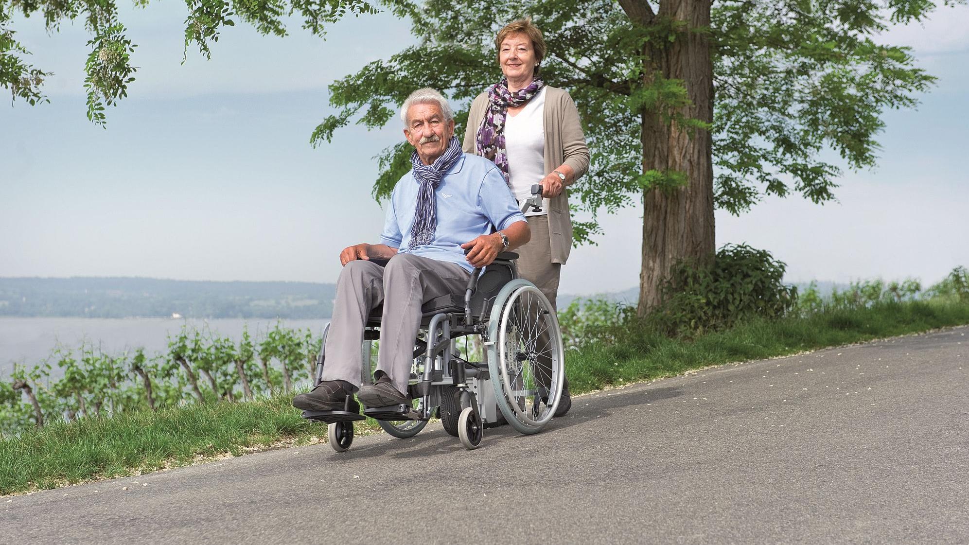 Ein im Rollstuhl sitzender Mann wird von einer Frau auf abschüssiger Straßegeschoben